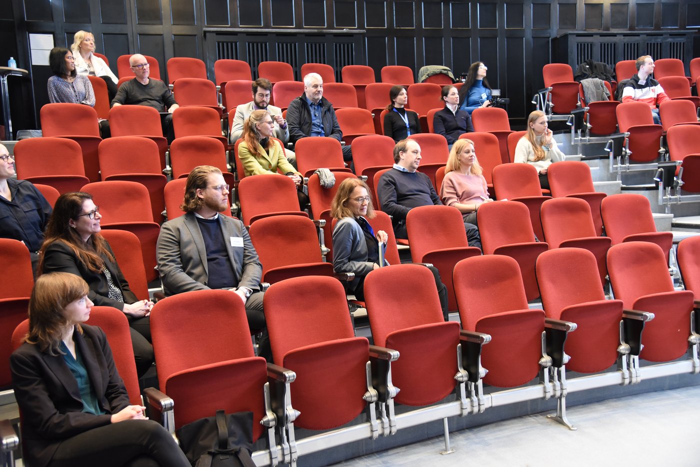 The picture shows the audience in the historic lecture hall on red upholstered folding seats in front of dark-panelled wooden walls.