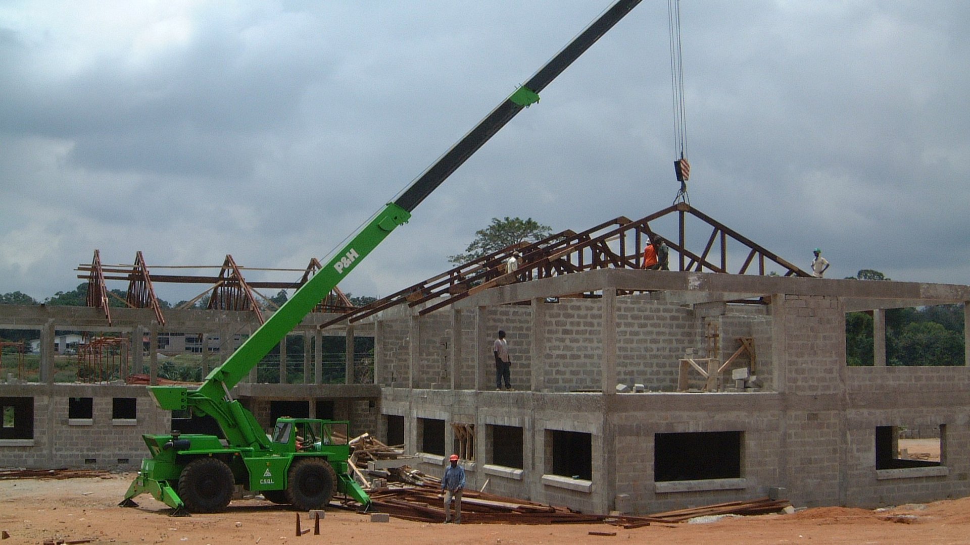 The picture shows a green crane in front of a house under construction with masonry and first roof beams.