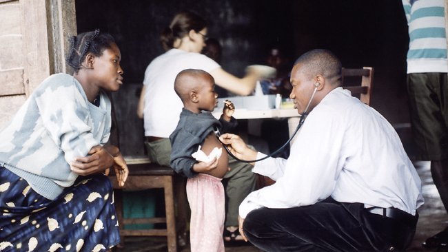 A Ghanaian doctor squats in front of a child, behind him his mother. The doctor examines the child with a stethoscope. In the background, a colleague from BNITM Hamburg with dark hair is working at a table.