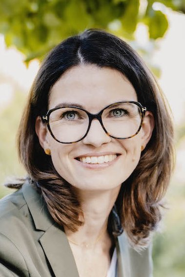 Portrait photo of Prof. Cornelia Betsch. A woman with shoulder-length brown hair and glasses is sitting under a tree with a very friendly smile. She is wearing an olive-green-grey suit with a white shirt underneath.