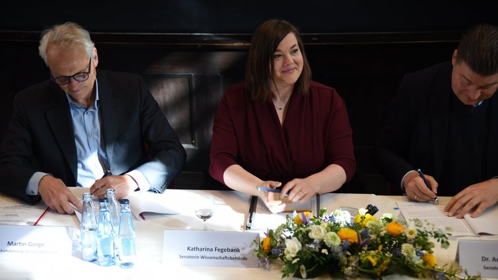 The picture shows from left to right Martin Görge (Sprinkenhof), Katharina Fegebank (Science Minister) and Dr Andreas Dressel (Finance Minister) signing the Letter of Intent. Ms Fegebank looks up with a smile while the others are still writing. In front of her is a colourful flower arrangement.
