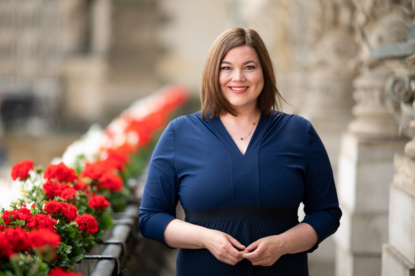 Katharina Fegebank: a politician on a balcony. On the left are red flowers, on the right the bright walls of Hamburg City Hall. Katarina Fegebank wears shoulder-length brown hair and a blue dress. In front of the middle of her body, the fingertips of her two hands are touching.