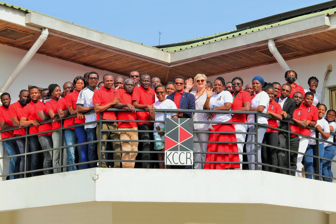 KCCR employees in predominantly red T-shirts stand on the balcony of the institute, some waving. At the front in the middle are the scientific director Prof. Phillips and the managing director Ingrid Sobel.