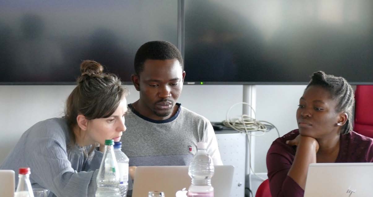 The photo shows three diverse people in the course room of the BNITM sitting in front of a teaching board on a laptop and discussing.